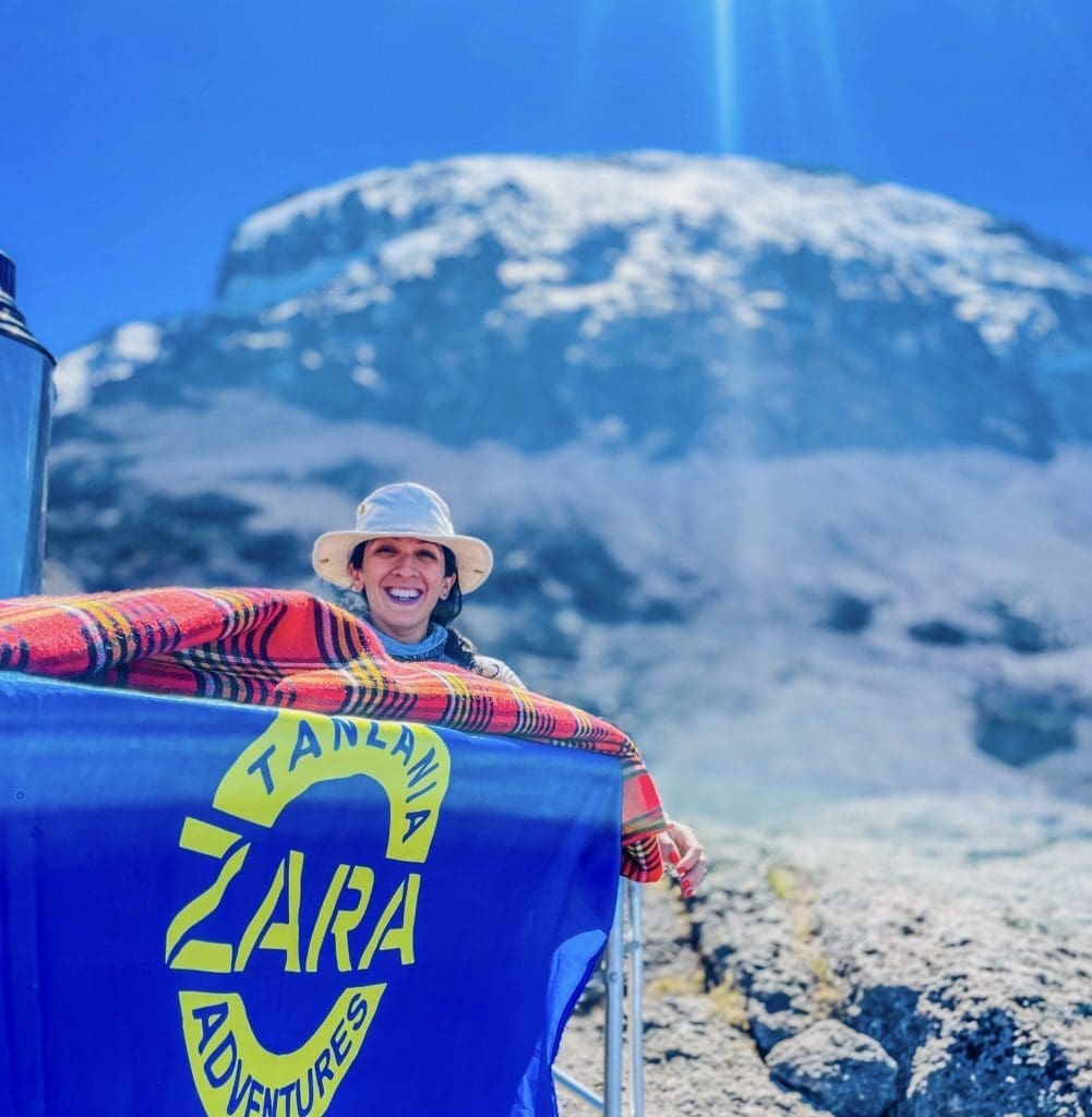 A smiling climber wrapped in a Maasai shuka holding a Zara Tanzania Adventures flag with snow-covered Mount Kilimanjaro in the background.