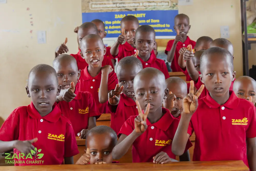Children wearing Zara Charity uniforms raise their hands in a classroom, representing the organization’s dedication to education and youth empowerment in Tanzania.