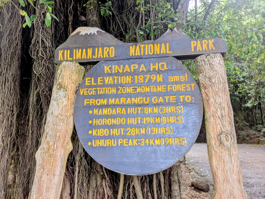 Wooden signboard at Kilimanjaro National Park (Kinapa HQ) showing elevation 1,879 meters and distances from Marangu Gate to Mandara Hut, Horombo Hut, Kibo Hut, and Uhuru Peak, surrounded by forest.