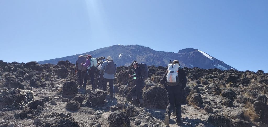 Group of climbers ascending a rocky alpine trail on Mount Kilimanjaro with backpacks, heading toward the summit under a clear blue sky.