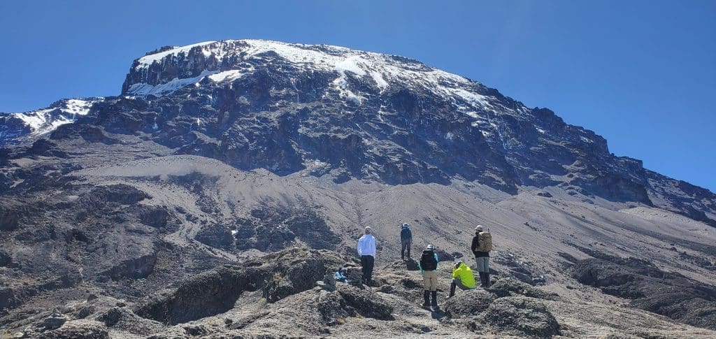 Climbers standing and resting on a rocky slope on Mount Kilimanjaro with the snow-capped summit rising above them under a clear blue sky.