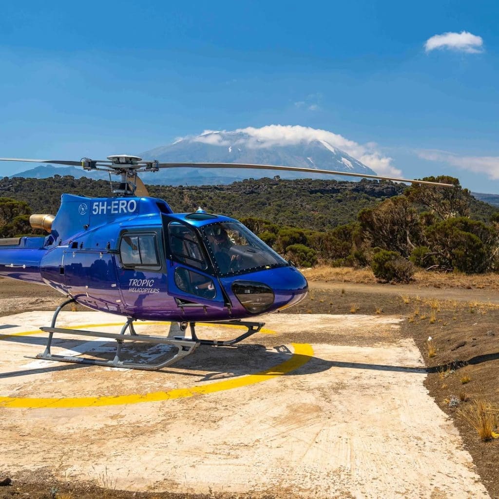 Blue rescue helicopter on a landing pad near Mount Kilimanjaro, with the snow-capped summit visible in the background.