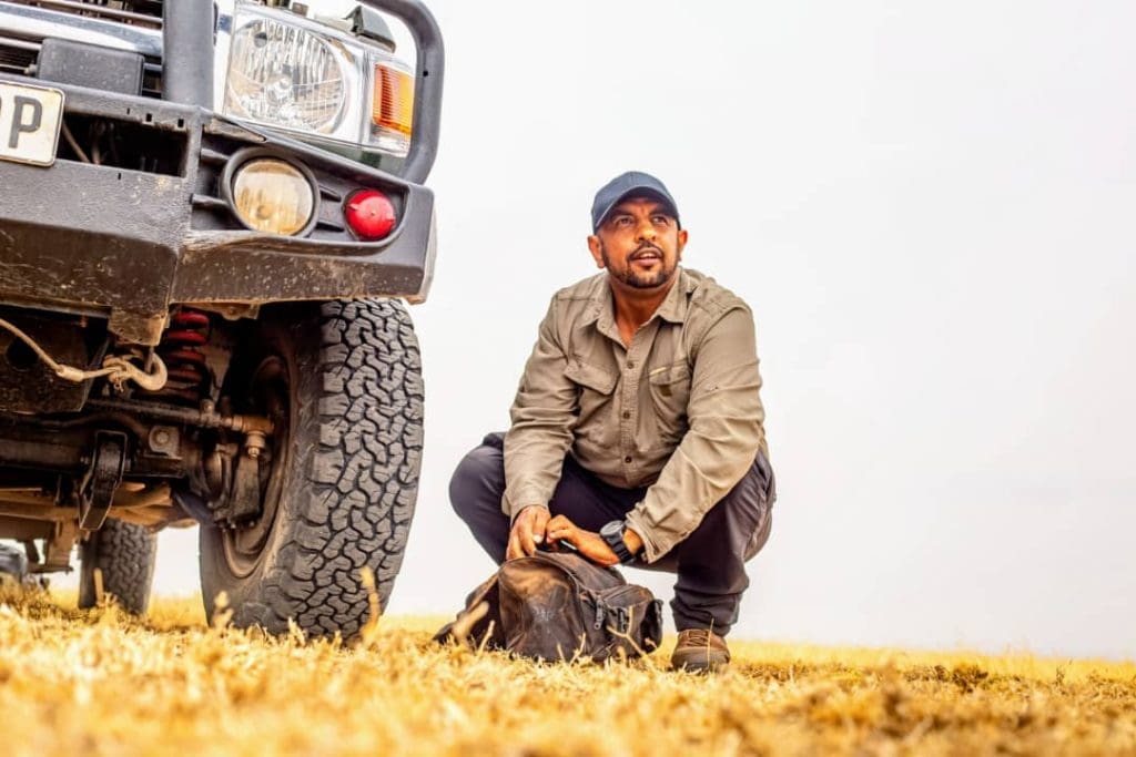Safari guide kneeling beside a 4x4 safari vehicle in the savannah, preparing gear for a game drive in Tanzania.