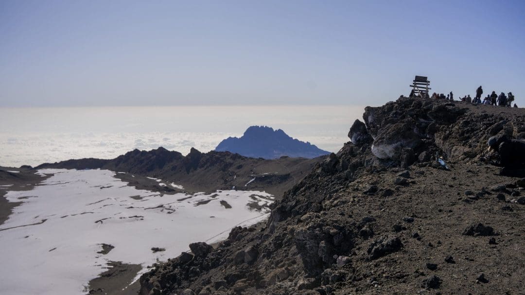 Snow-covered slopes and rocky terrain near the summit of Mount Kilimanjaro, showing changing high-altitude weather conditions above the cloud layer.