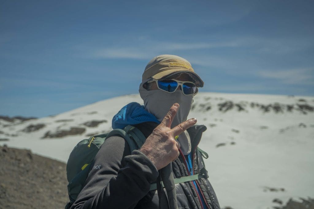 Climber wearing sunglasses, face covering, and warm layers on the snowy slopes of Mount Kilimanjaro to protect against strong sun, cold wind, and high-altitude conditions.