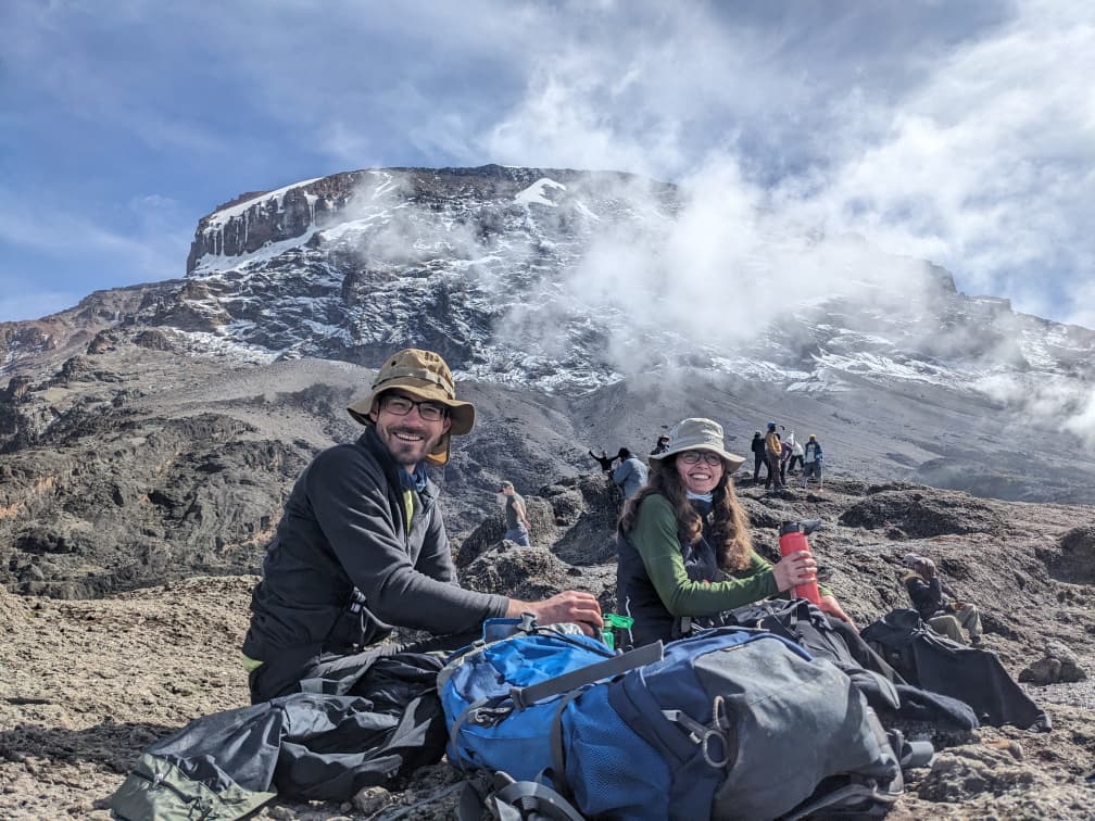 Two climbers resting on rocky terrain high on Mount Kilimanjaro with backpacks, clouds drifting around the summit behind them.
